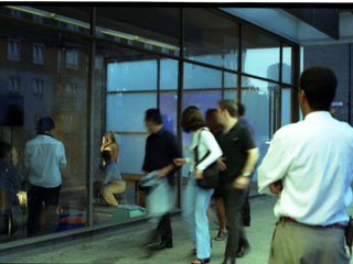 A still image from the performance In 24 Hours by Corali Dance Company, 2000. The photo shows the back the front of the empty shop unit with members of the public walking past the window looking in through the windows at the live action of the performance happening on the inside of the shop window.